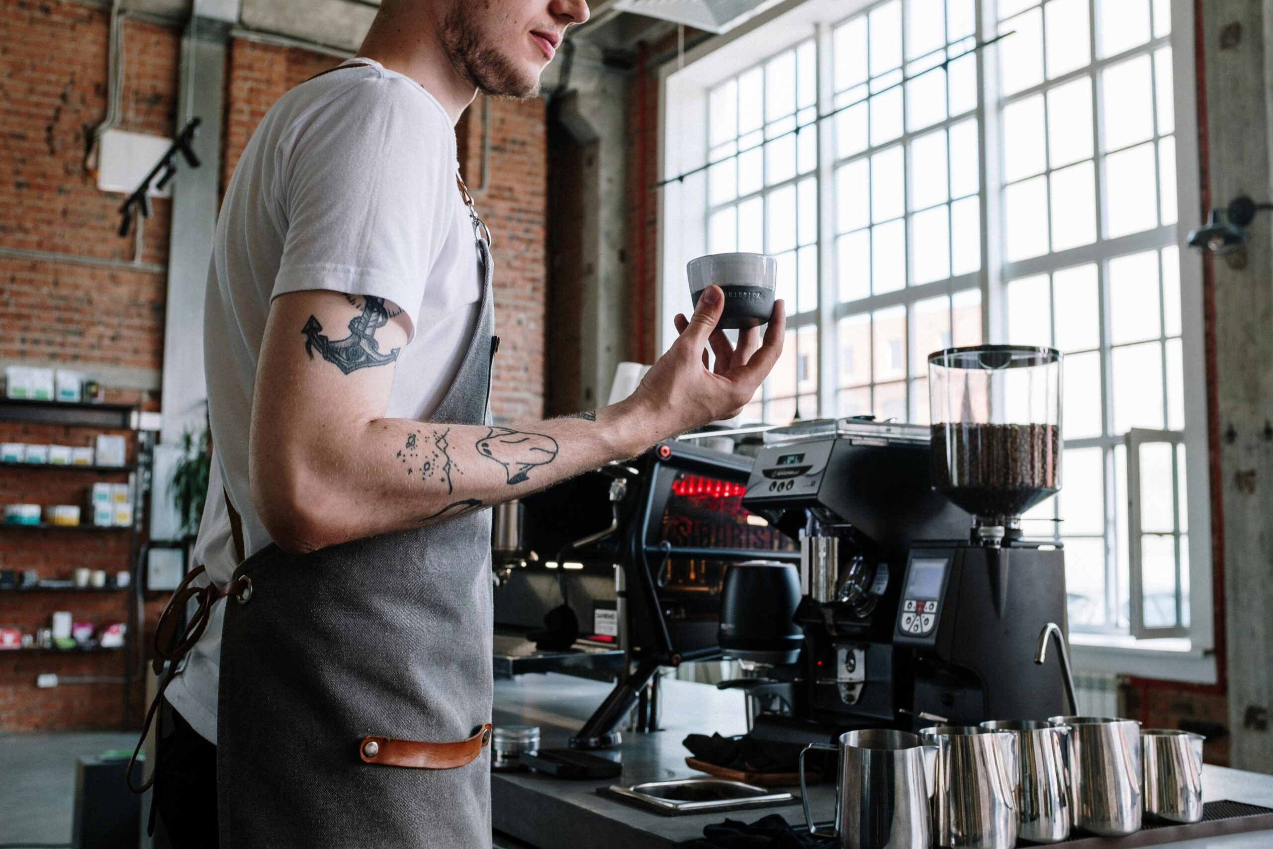 A professionally trained barista with tattoos holds an espresso cup behind an espresso machine and grinder