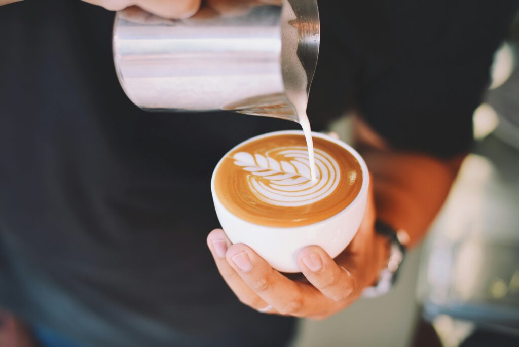 A barista is pouring steamed milk from a metal pitcher into a latte creating art